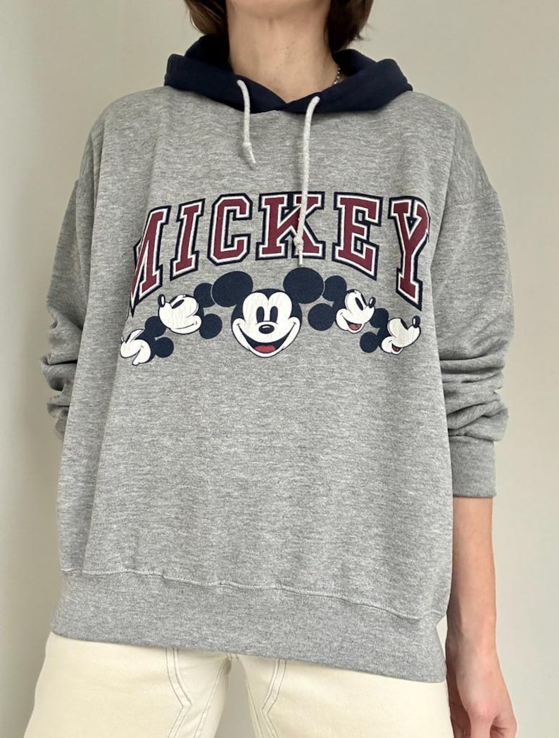 May include: Gray sweatshirt with a navy blue hood and white drawstring. The word "MICKEY" is printed across the chest in maroon and white, with Mickey Mouse graphics below. The sweatshirt is a relaxed fit.