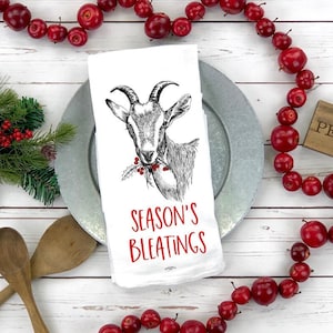 Könnte beinhalten: Ein weißes Küchentuch mit einer Schwarzweißillustration einer Ziege mit dem Text "Season's Bleatings" in Rot. Das Tuch liegt auf einem silbernen Teller mit einer roten Beerenkette drumherum.