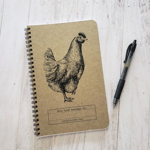 Puede incluir: Una libreta de espiral marrón con una ilustración en blanco y negro de una gallina en la portada. La libreta tiene el texto "this book belongs to:" y "(crazy chicken lady!)" impreso en la portada.