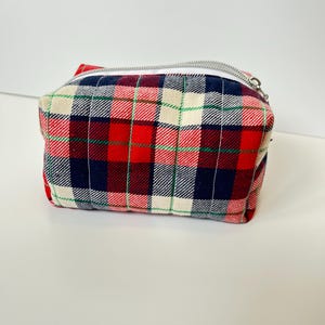Peut inclure: Une petite pochette rectangulaire à carreaux avec une fermeture éclair argentée. Le tissu présente un motif à carreaux classique rouge, marine, blanc et vert. La pochette est probablement utilisée pour les cosmétiques ou les petits objets.