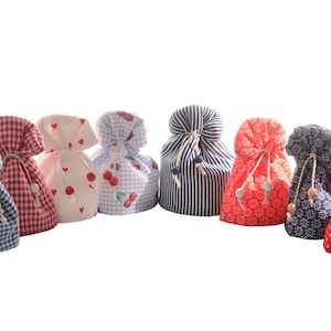 Könnte beinhalten: Eine Sammlung von acht Stoffbeuteln mit Kordelzug in verschiedenen Mustern und Farben. Designs umfassen Vichy, Streifen, Kirschen, Herzen und Blumendrucke. Jeder Beutel ist mit einem Kordelzug und Holzperlen gebunden.