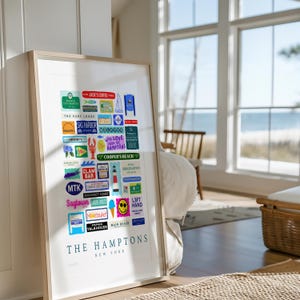 May include: Framed art print featuring a collage of colorful signs and labels from The Hamptons, New York. The print includes various businesses and landmarks, with text such as "Jack's Coffee", "Sag Harbor", and "Cooper's Beach". The title "The Hamptons" is displayed at the bottom.
