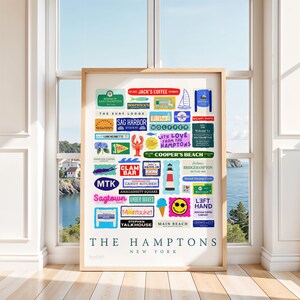 May include: A framed print featuring colorful signs for various locations in The Hamptons, New York. The signs include names like Sag Harbor, Montauk, and Main Beach, with a backdrop of a coastal scene visible through a window.