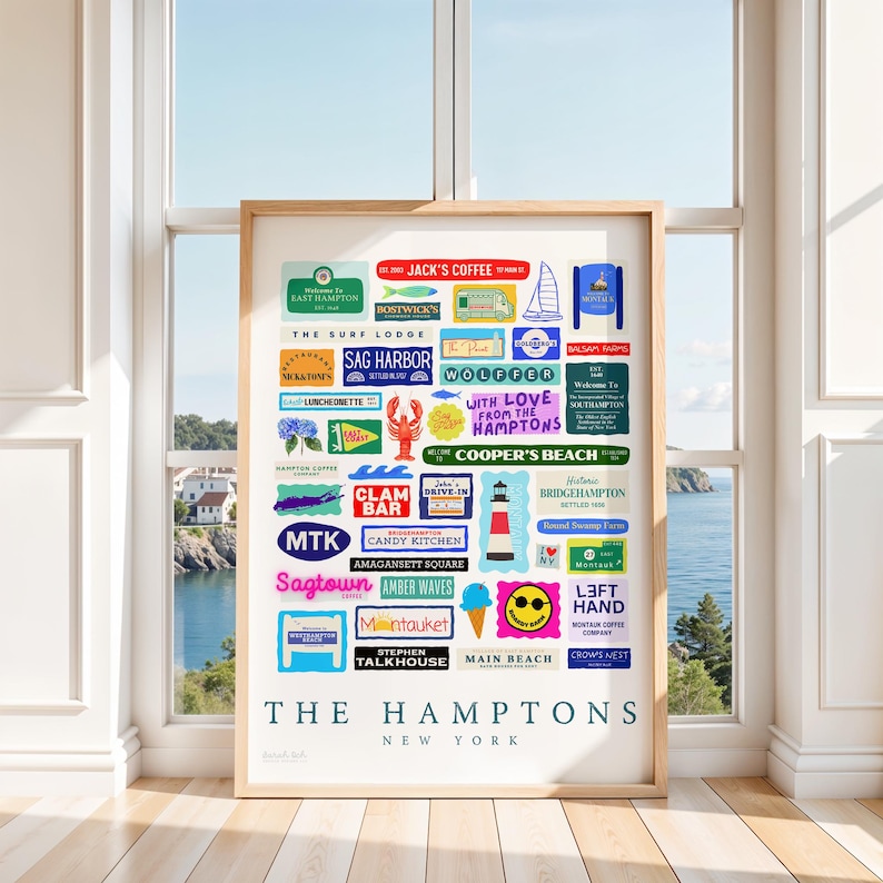May include: A framed print featuring colorful signs for various locations in The Hamptons, New York. The signs include names like Sag Harbor, Montauk, and Main Beach, with a backdrop of a coastal scene visible through a window.