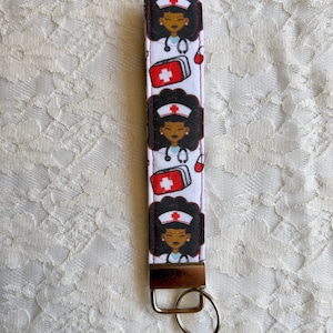 May include: White keychain with a repeating pattern of a cartoon Black nurse wearing a white uniform and a red cross. The nurse is holding a red first aid kit. The keychain has a silver key ring.