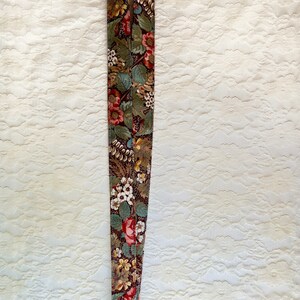 Puede incluir: Un cordón con estampado floral y un llavero de metal dorado. El cordón es marrón con flores rojas, verdes y blancas.