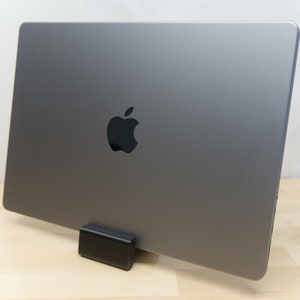 May include: A silver laptop computer with an Apple logo on the back is sitting on a black stand. The laptop is on a light wood surface.