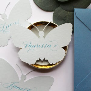 May include: White butterfly-shaped place cards with teal calligraphy names, one reading "Marissa." The cards sit on a gold-rimmed dish, with a blue envelope and eucalyptus leaves in the background. A delicate and elegant design.