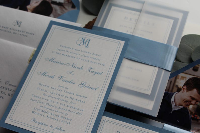 May include: Elegant wedding invitations with a light blue border and matching ribbon. The invitation includes the names of the couple, date, and location of the ceremony. The design features a monogram and detailed text.