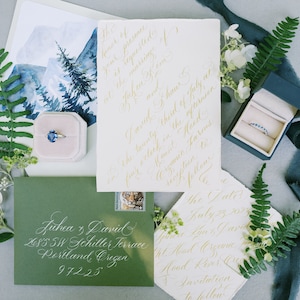 May include: A wedding invitation suite with a green envelope, a white card with gold calligraphy, a blue ring box, and a watercolor painting of a mountain landscape. The invitation text reads "The Late July 23, 2023 at Mead Oregon. Invitation to follow."