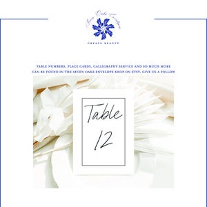 May include: A white table number card with the number 12 in black script, framed by a thin black border. The card is set against a backdrop of white flowers and soft, neutral tones. The top of the image features a blue floral logo and the words "CREATE BEAUTY".
