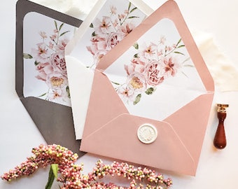 Pode incluir: Três envelopes com desenhos florais. Os envelopes são em tons de rosa, cinza e branco. Os envelopes são decorados com um selo de cera e um desenho floral. Os envelopes estão sobre uma superfície branca.