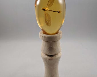 May include: A clear amber stone with a preserved dragonfly inside, displayed on a wooden stand.