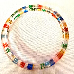 May include: A clear, circular bracelet with colorful puzzle piece accents. The bracelet features red, blue, green, yellow, and orange puzzle pieces embedded within the clear material. The bracelet is a unique accessory.