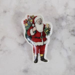 May include: A ceramic ornament featuring a cartoon image of Santa Claus carrying a bag of gifts. Santa is wearing a red suit with a white trim, black boots, and a white beard.