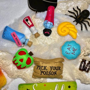 May include: A collection of felt crafts inspired by classic fairy tales. The crafts include a poison apple, a bottle labeled "Drink Me", a scroll that says "Pick Your Poison", a cookie that says "Eat Me", a seashell, a bottle of potion, and a spider.