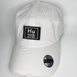 Könnte beinhalten: Eine weiße Baseballkappe mit Used-Optik und einem rechteckigen schwarzen Aufnäher auf der Vorderseite mit dem weißen Text Hu Hustle 24.7.365. Ein runder holografischer Aufkleber mit dem Text KBETHOS Vintage SINCE 2001 befindet sich auf dem Schirm.