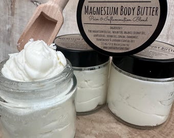 May include: Three glass jars filled with a white cream labeled "Magnesium Body Butter Pain & Inflammation Blend" with the brand name "Qadash Creations". The jars are on a wooden surface with a wooden scoop.