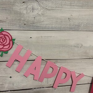 May include: Pink paper banner with the words "Happy" and a rose decoration. The banner is strung with white twine and is hanging on a whitewashed wood background.