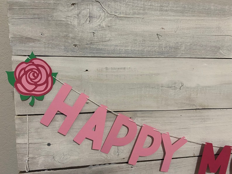 May include: Pink paper banner with the words "Happy" and a rose decoration. The banner is strung with white twine and is hanging on a whitewashed wood background.