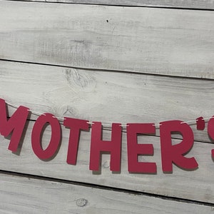 May include: Red felt banner with the word "MOTHER'S" strung on white twine against a light wood background.