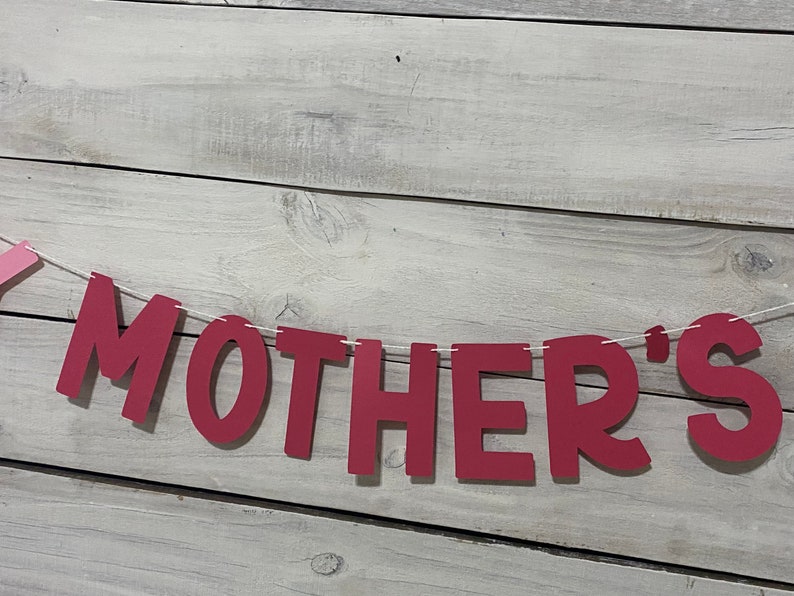 May include: Red felt banner with the word "MOTHER'S" strung on white twine against a light wood background.
