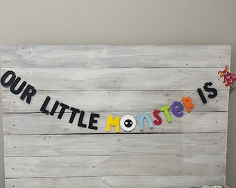 Puede incluir: Una pancarta colorida con las palabras "Our Little Monster Is" en letras negras, amarillas, blancas, azules, rojas y moradas. Un pequeño monstruo rojo con tres ojos está en el lado derecho de la pancarta.