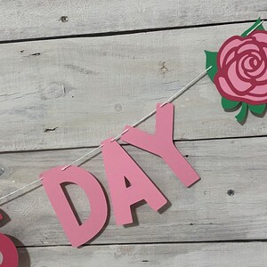 May include: Pink paper banner with the word "DAY" in large letters and a pink rose with green leaves. The banner is strung with white twine.