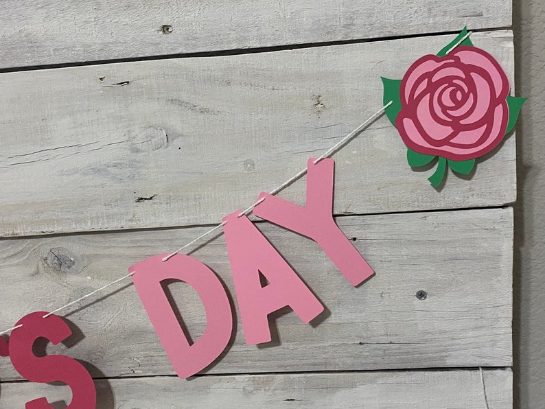 May include: Pink paper banner with the word "DAY" in large letters and a pink rose with green leaves. The banner is strung with white twine.