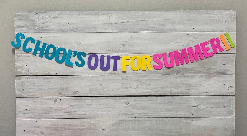 May include: A colourful paper banner with the text "SCHOOL'S OUT FOR SUMMER" hanging on a whitewashed wood background.