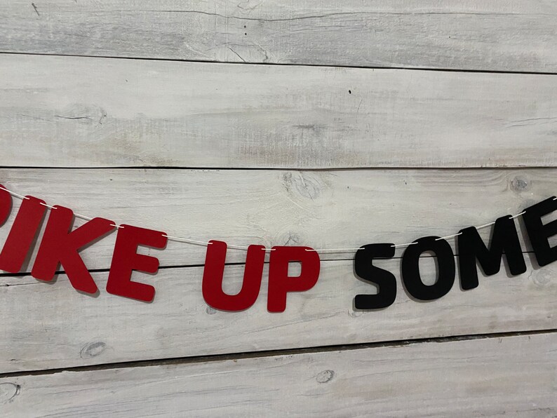 May include: Red and black felt letter banner that reads "Pike Up Some". The banner is strung on a white string and is hanging against a white wood background.