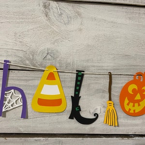 May include: A Halloween banner with the word "HALLOWEEN" spelled out in paper cutouts. The letters are decorated with various Halloween themes, including a spiderweb, a candy corn, a witch's boot, a broom, and a jack-o'-lantern.