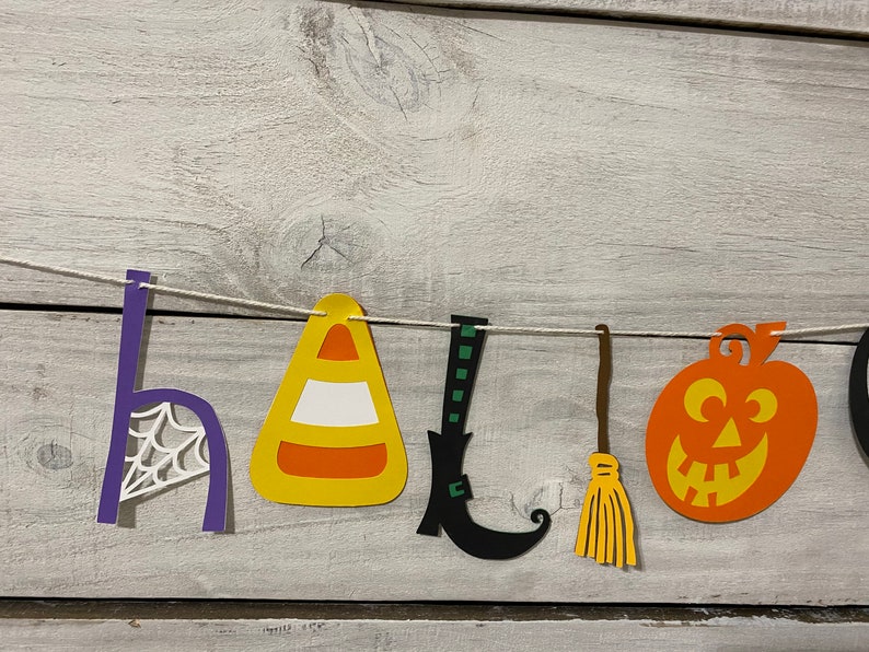 May include: A Halloween banner with the word "HALLOWEEN" spelled out in paper cutouts. The letters are decorated with various Halloween themes, including a spiderweb, a candy corn, a witch's boot, a broom, and a jack-o'-lantern.