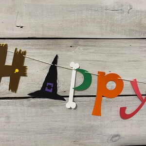 May include: A Halloween banner with the word "Happy" spelled out in colorful paper letters. The "H" is brown with yellow accents, the "a" is a witch's hat in black with purple accents, the "p" is a white bone, the "p" is green, the "y" is orange, and the "y" is red.