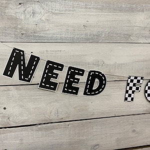 Op de afbeelding: Zwart-witte banner met de woorden "NEED FOR" en een geruit patroon. De banner is aan een wit touw bevestigd en hangt aan een witte houten muur.