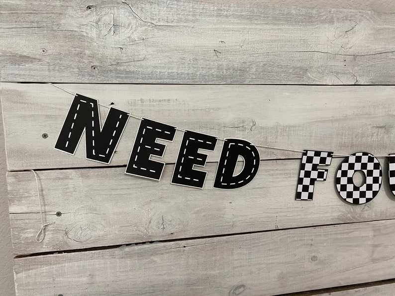 Op de afbeelding: Zwart-witte banner met de woorden "NEED FOR" en een geruit patroon. De banner is aan een wit touw bevestigd en hangt aan een witte houten muur.