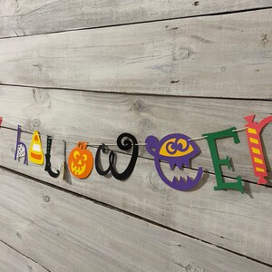 May include: A colorful paper banner with the words "Happy Halloween" in different colors and styles. The banner includes a witch's hat, a spider web, a candy corn, a broom, a pumpkin, and a monster.