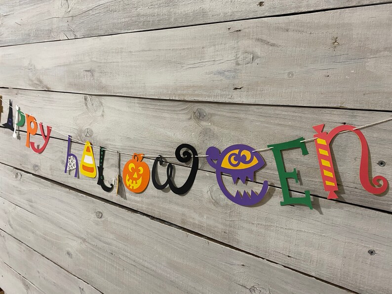May include: A colorful paper banner with the words "Happy Halloween" in different colors and styles. The banner includes a witch's hat, a spider web, a candy corn, a broom, a pumpkin, and a monster.