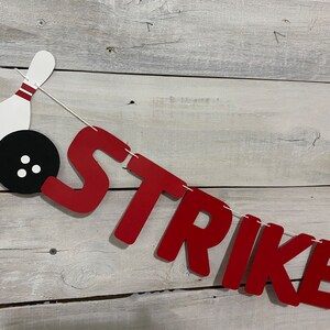 May include: Red felt banner with the word "STRIKE" in white letters. A white bowling pin with red stripes and a black bowling ball with white dots are attached to the banner.