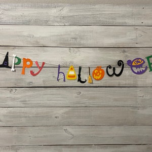 May include: A colorful Happy Halloween banner with cut-out letters and festive Halloween icons, including a witch's hat, a candy corn, a pumpkin, a broom, and a ghost. The banner is strung on a white background.