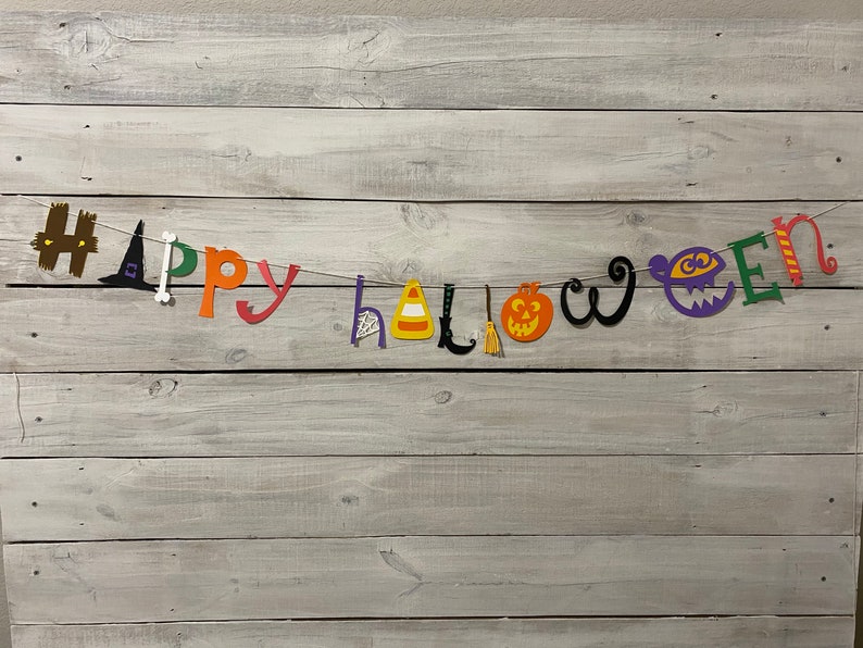 May include: A colorful Happy Halloween banner with cut-out letters and festive Halloween icons, including a witch's hat, a candy corn, a pumpkin, a broom, and a ghost. The banner is strung on a white background.