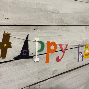 May include: A paper banner with the words "Happy Halloween" in colorful letters and cut-out Halloween icons. The banner is strung on a white string and is hanging on a white wooden wall.
