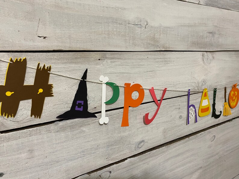May include: A paper banner with the words "Happy Halloween" in colorful letters and cut-out Halloween icons. The banner is strung on a white string and is hanging on a white wooden wall.