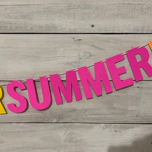 May include: A colourful paper banner with the words "FOR SUMMER" in pink and yellow letters. The banner is strung on a white string and is hanging against a white wood background.