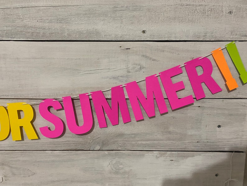 May include: A colourful paper banner with the words "FOR SUMMER" in pink and yellow letters. The banner is strung on a white string and is hanging against a white wood background.