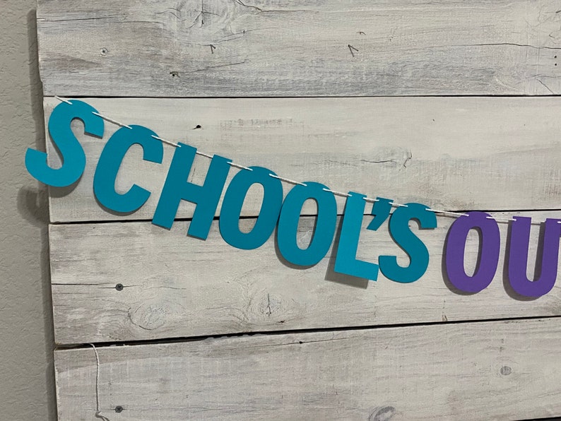 May include: A teal blue and purple paper banner with the text "SCHOOL'S OUT" hanging on a white wood background.