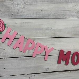 May include: Pink and red paper banner with the words "Happy Mother's" hanging on a white wood background. A pink paper rose with green leaves is at the top left of the banner.