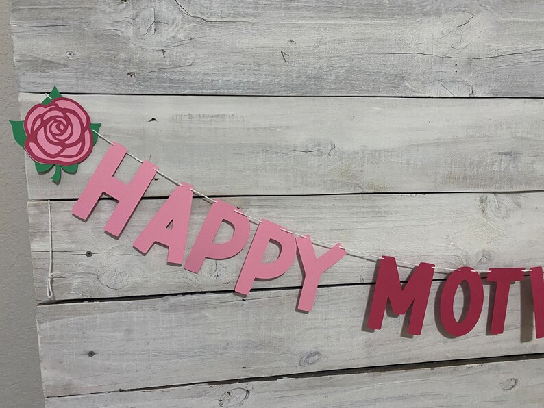 May include: Pink and red paper banner with the words "Happy Mother's" hanging on a white wood background. A pink paper rose with green leaves is at the top left of the banner.