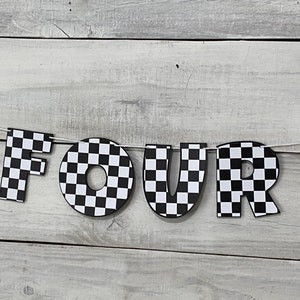 Op de afbeelding: Zwart-wit geblokt spandoek met het woord "VIER" in grote letters. Het spandoek is aan een wit touw bevestigd en hangt tegen een witte houten achtergrond.