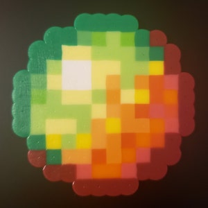 May include: A pixel art design made with colorful beads, featuring a green, yellow, orange, and red color scheme. The design is circular and resembles a pixelated image.